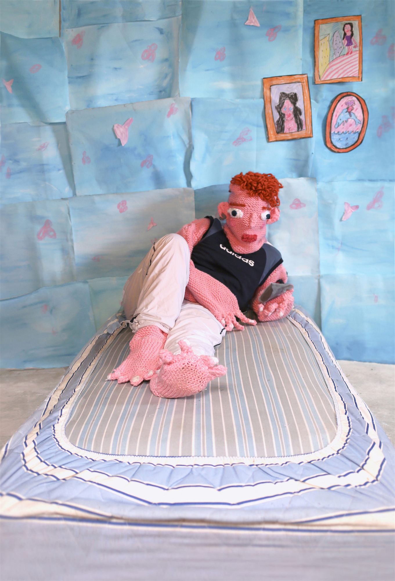 Éva Ostrowska, Jason waiting for her text, pink knitted figure in an Adidas top on a bed against a hand-painted blue paper room, photographic performance