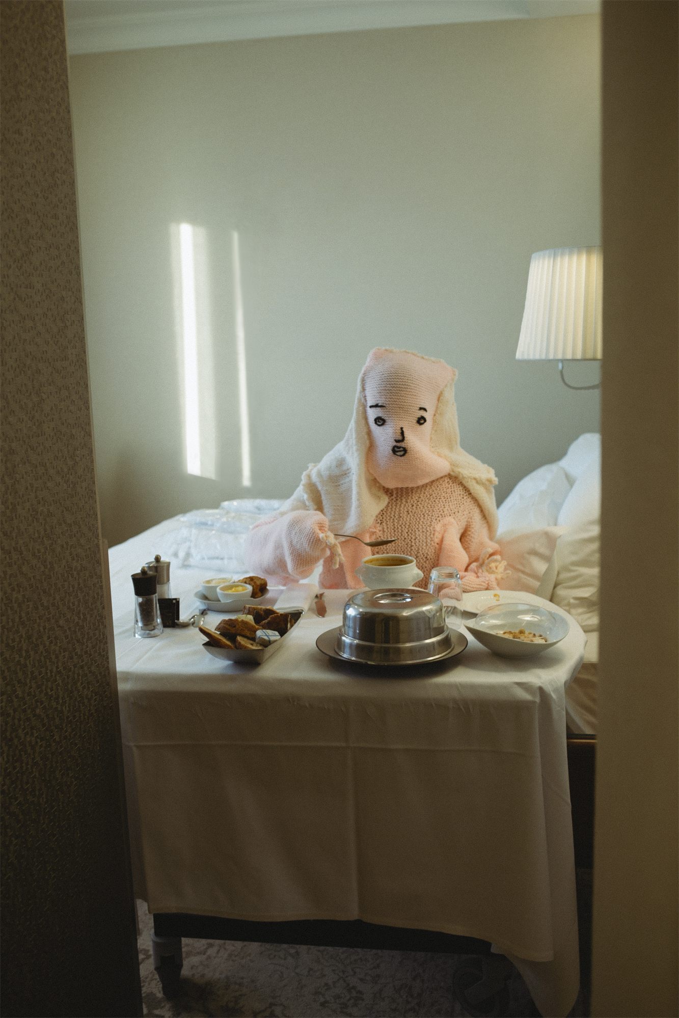 Éva Ostrowska, Le Souper, knitted figure taking room service dinner in bed in a cream hotel room, photographic performance