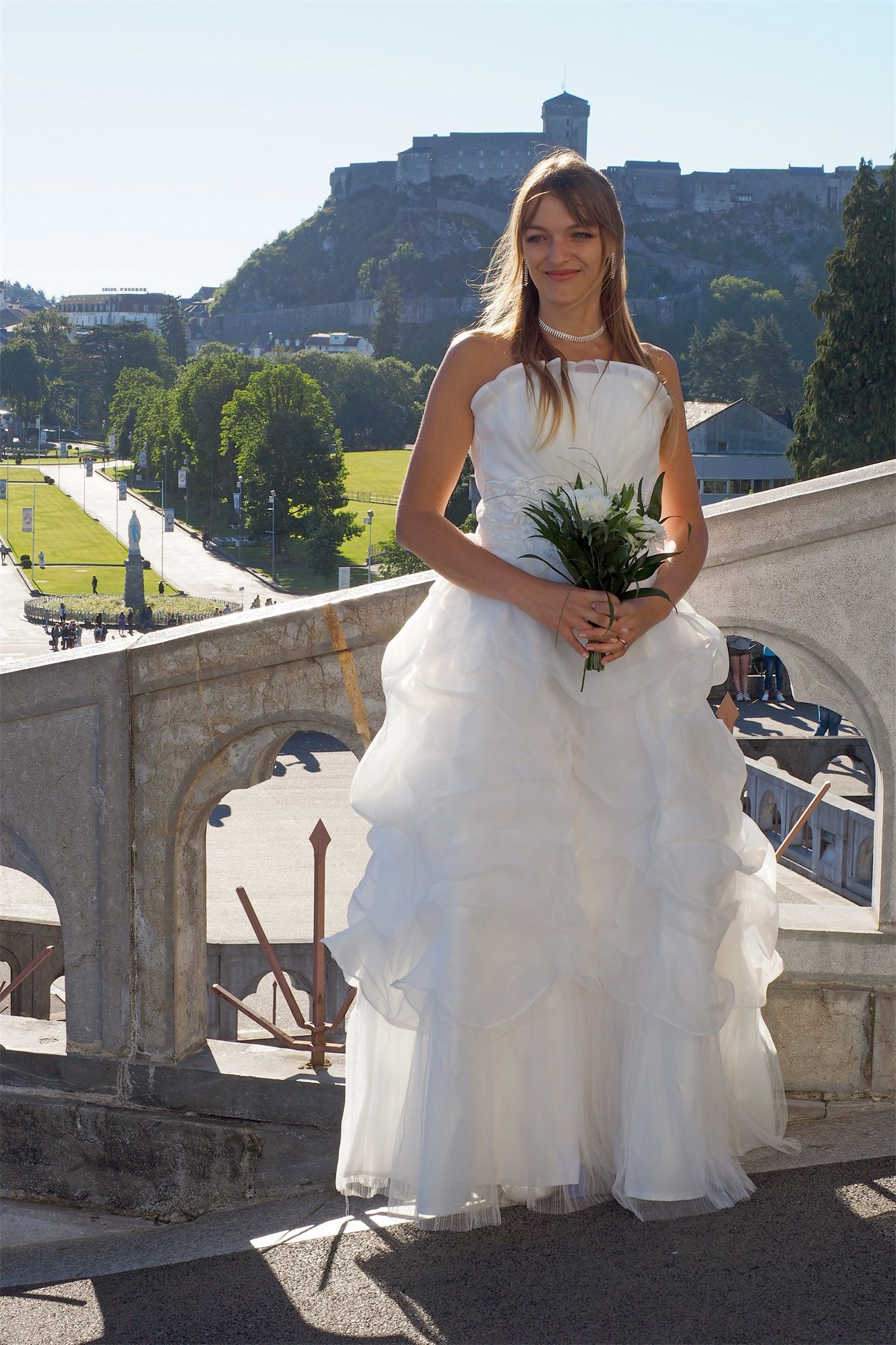 Éva Ostrowska in a wedding dress on the terrace of the Rosary Basilica, the castle of Lourdes behind, 2016