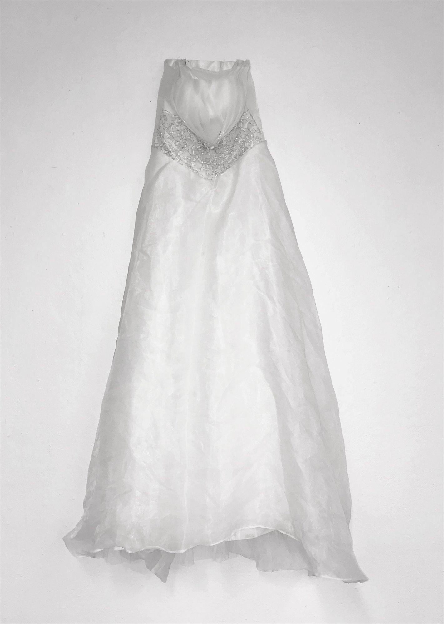 The white organza wedding dress from the 2016 self-marriage of Éva Ostrowska, hung empty on a white wall